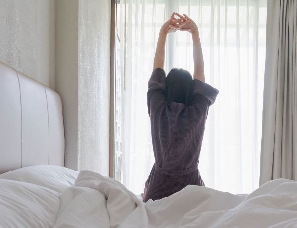 Frau sitzt im Bett und streckt beim Aufwachen die Arme nach oben.