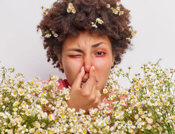 Person mit lockigem Haar hält sich die Nase zu, umgeben von kleinen weißen Blumen.