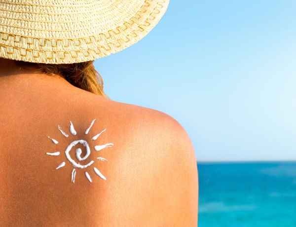 Frau mit Sonnenhut sitzt am Strand, auf der Schulter ist ein Sonne-Symbol aus Sonnencreme aufgetragen.