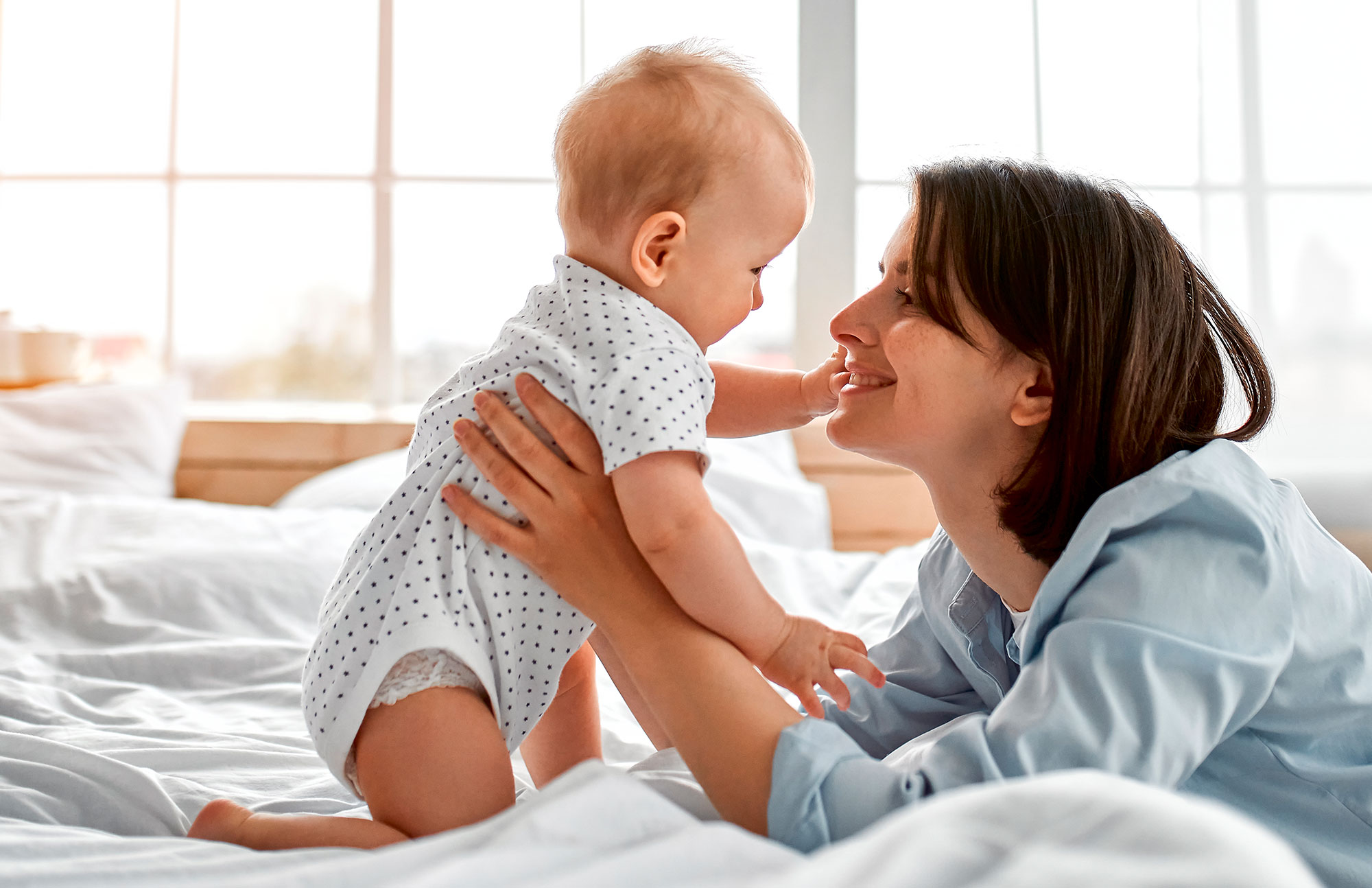 Mutter spielt im Bett mit ihrem Baby, beide lachen fröhlich.