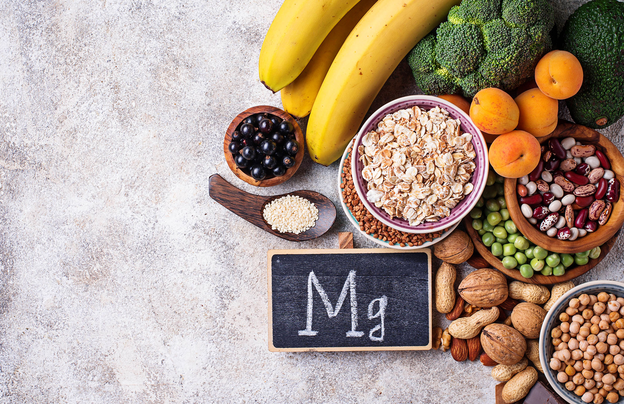 Verschiedene magnesiumreiche Lebensmittel wie Nüsse, Bananen, Kakao und Trockenfrüchte auf grauem Untergrund.
