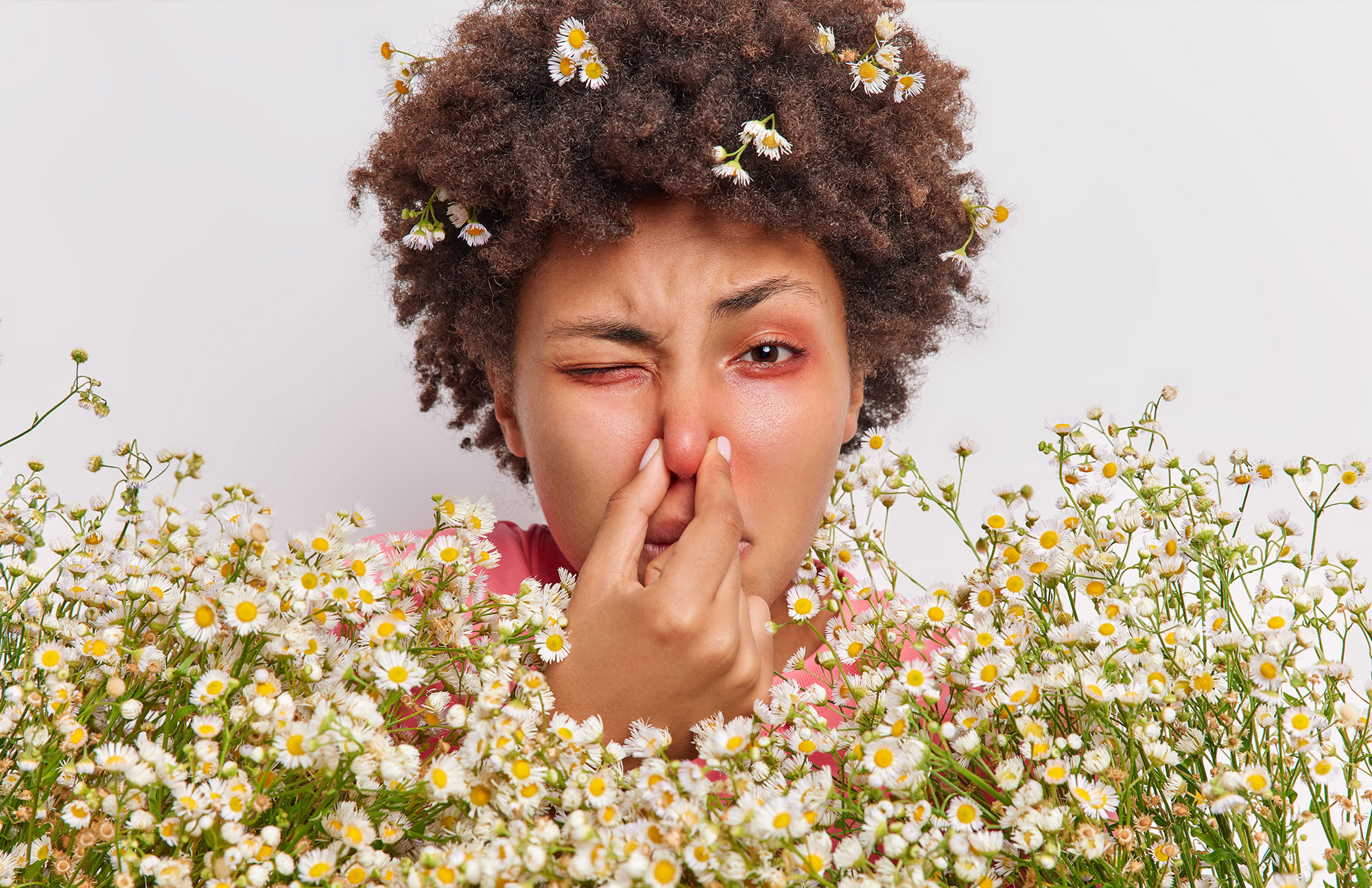 Person mit lockigem Haar hält sich die Nase zu, umgeben von kleinen weißen Blumen.