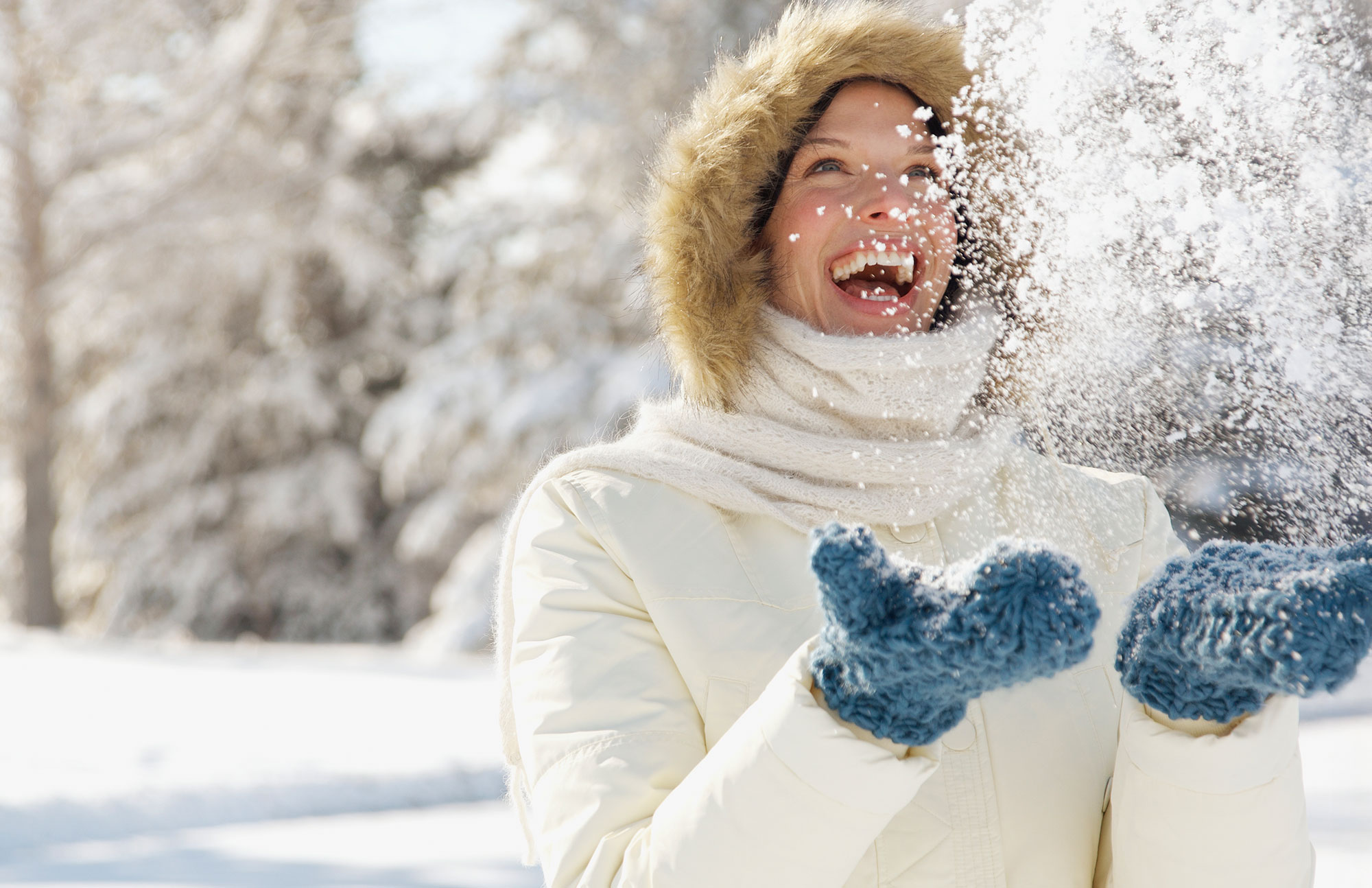 Frau in Winterjacke und Handschuhen wirft lachend Schnee in die Luft.