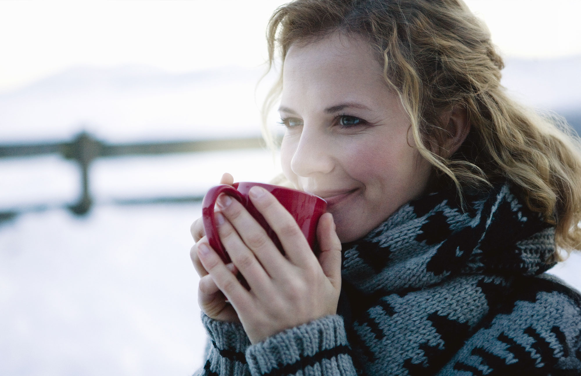 Frau mit Schal hält eine rote Tasse in den Händen, winterliche Landschaft im Hintergrund.